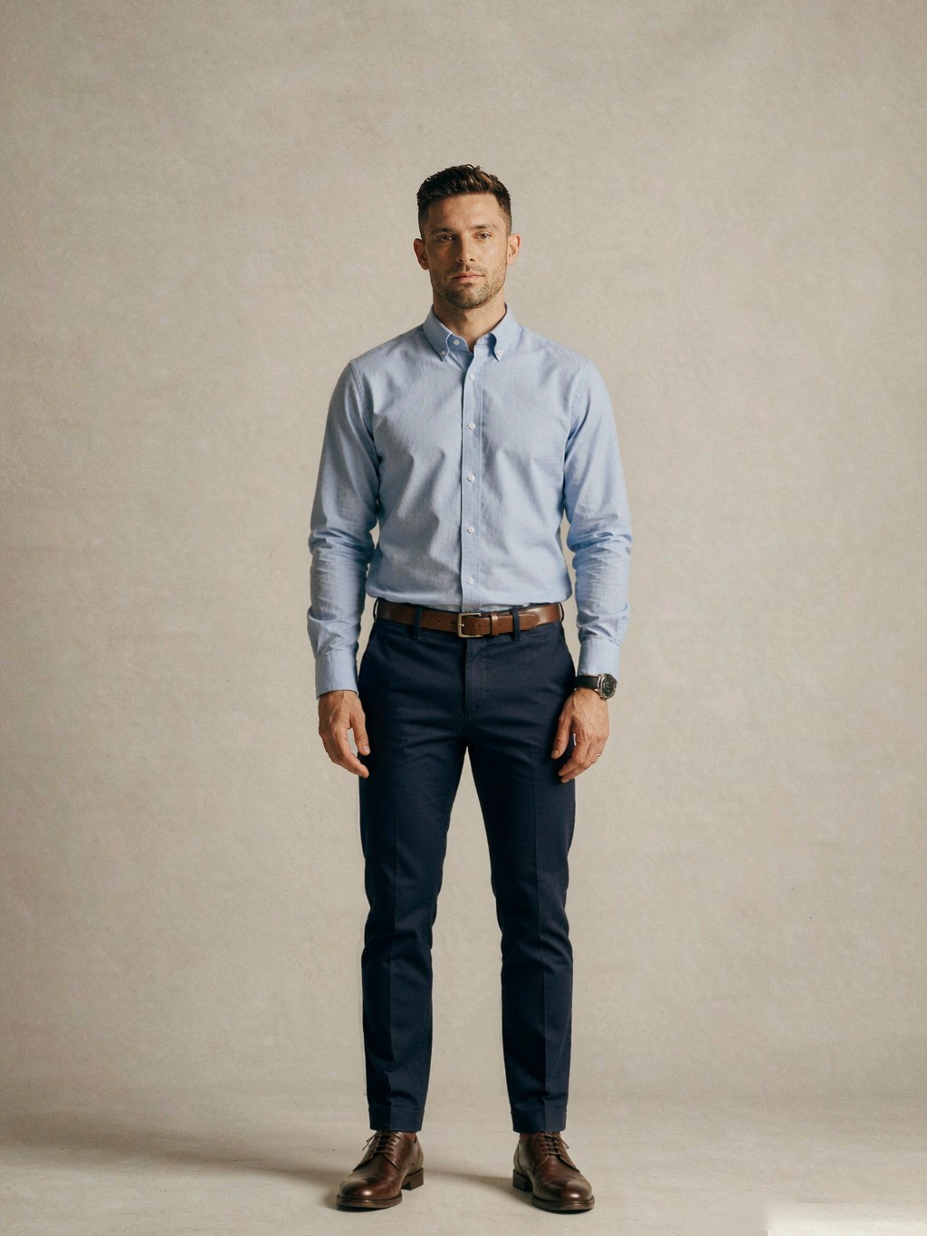 Full body, front view: light blue button-down oxford shirt tucked into straight-leg navy chinos with a brown leather belt and dark brown leather derby shoes